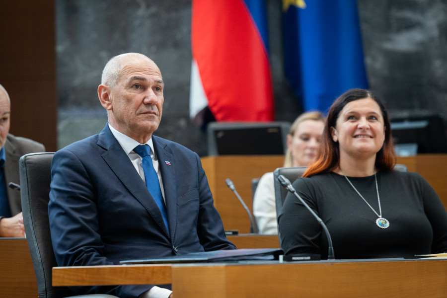 A man in a blue suit and a woman wearing a pendant sit at a conference table beneath national and EU flags.