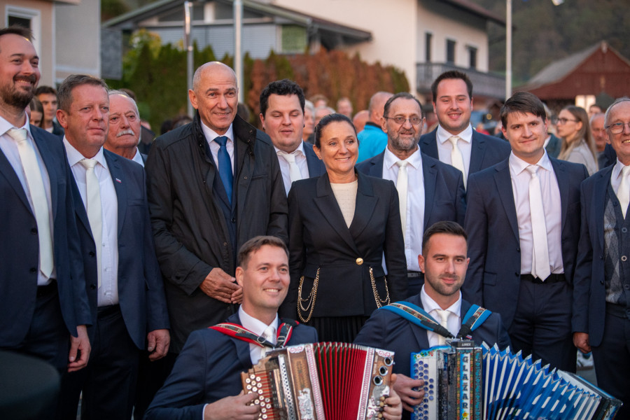 A formally dressed group poses outdoors behind two seated musicians holding accordions at a public gathering.