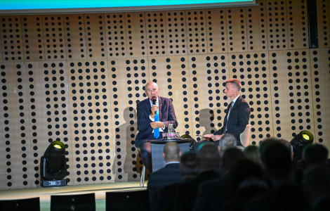 MK1 1538 (Two suited men discuss a topic onstage, one speaking into a microphone as an audience watches from the foreground.)