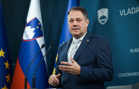 nk rezultati dela vlade arcon (The man is standing behind a podium; behind him on the wall are the Slovenian coat of arms and the word “government,” and to his left is the Slovenian flag.)