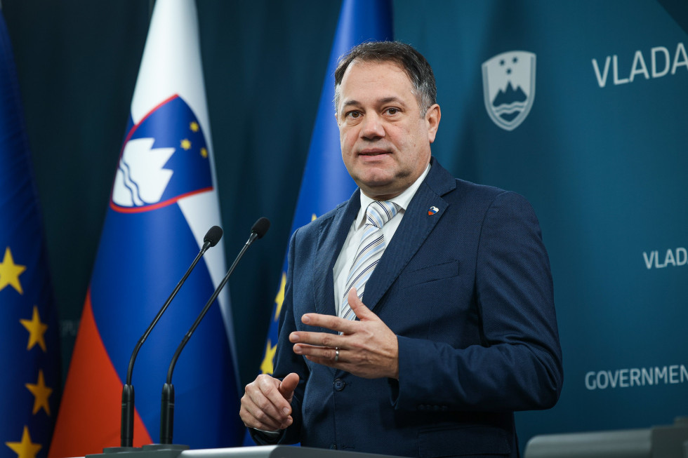 Moški stoji za govorniškim pultom, za njim na steni slovenski grb in napis vlada, levo od njega slovenska zastava. 