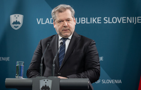 papic (A man in a dark suit speaking at a press conference, with the logo and name of the Government of the Republic of Slovenia in the background.)