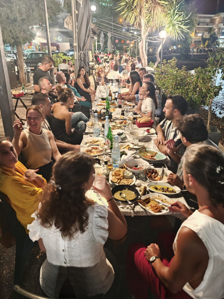 Okoli velike, s hrano obložene mize sedijo moški in ženske, jedo in govorijo.