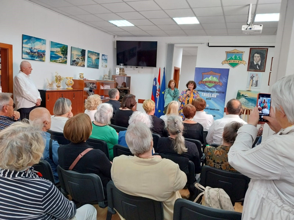 V dvorani sedijo poslušalci, pred njimi stoji ženska v pisani bluzi in govori na mikrofon. Za njo je plakat z napisom Slovensko kulturno društvo Triglav Split.