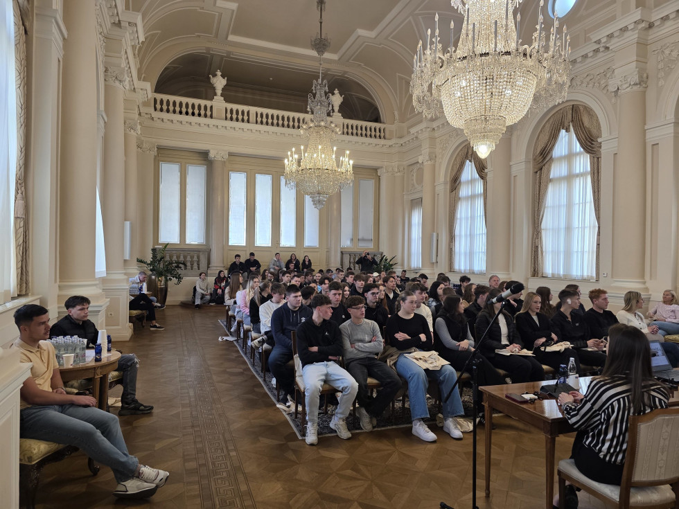 Skupina dijakov sedi na stolih v kristalni dvorani in posluša.