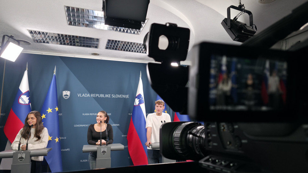 Tri osebe stojijo za govorniškimi pulti na tiskovni konferenci, za njimi pa so zastave Slovenije in Evropske unije. V ospredju je profesionalna videokamera, ki zamegli subjekte in njihove posamezne izraze.