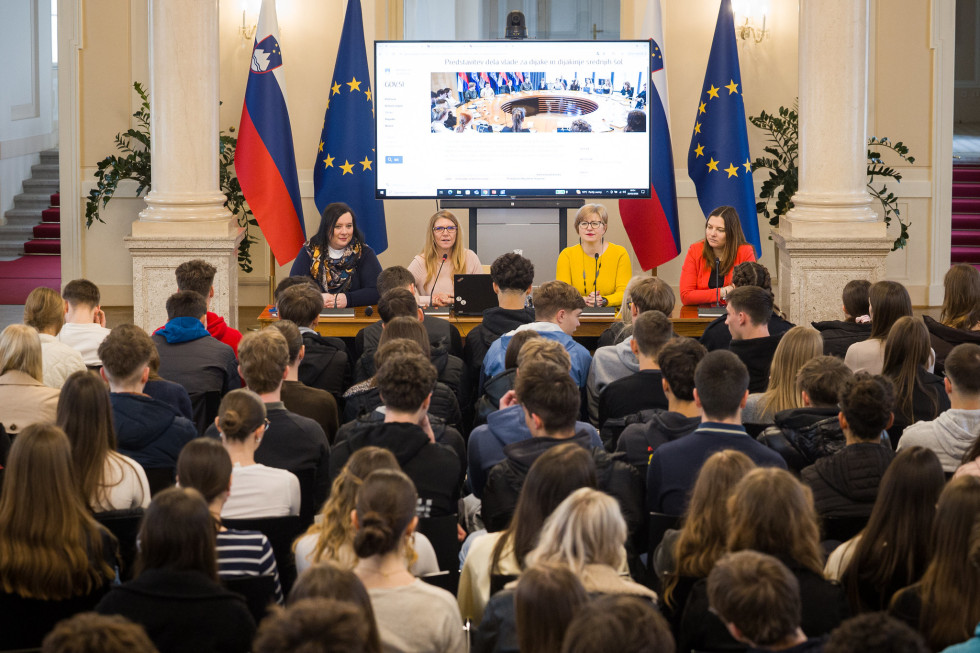 Dvorana polna mladih dijakov, ki sedijo in gledajo proti govorkam. Za govorniško mizo sedijo štiri ženske, za njimi večji ekran.