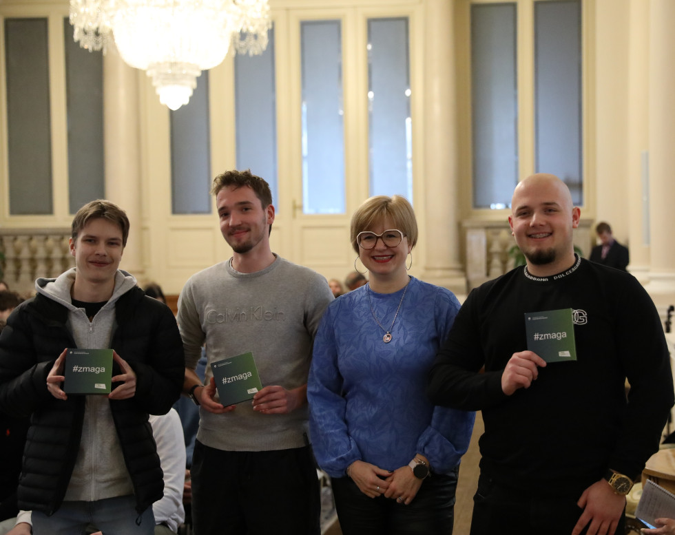Trije mlajši moški in ženska stojijo in pozirajo. Moški v rokah držijo manjšo škatlico z napisom #zmaga.