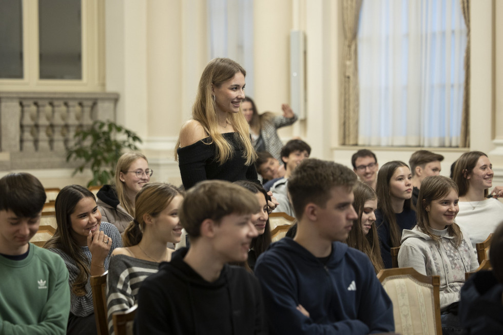 Mlada ženska stoji, ostali mladi okoli nje sedijo in se smejijo.
