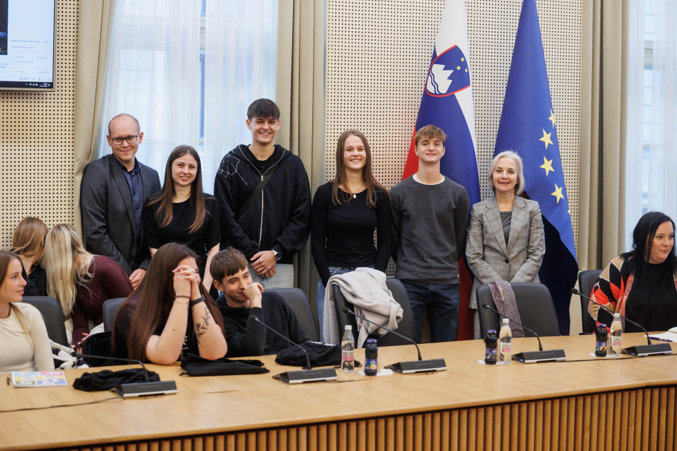 Skupina odraslih in najstnikov pozira za konferenčno mizo v prostoru. Za njimi sta slovenska in EU zastava. Osebe so oblečene v različnih kombinacijah sproščenih in formalnih oblačil, njihove izraze pa večinoma odlikuje nevtralen ali rahlo nasmejan izraz.