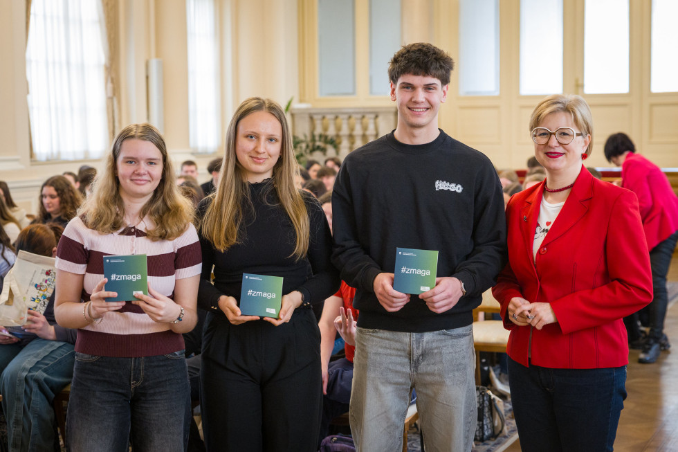 Trije mladi pozirajo fotografu, v rokah držijo škatlice z napisom #zmagovalci. Desno ob njih ženska v rdečem blazerju.