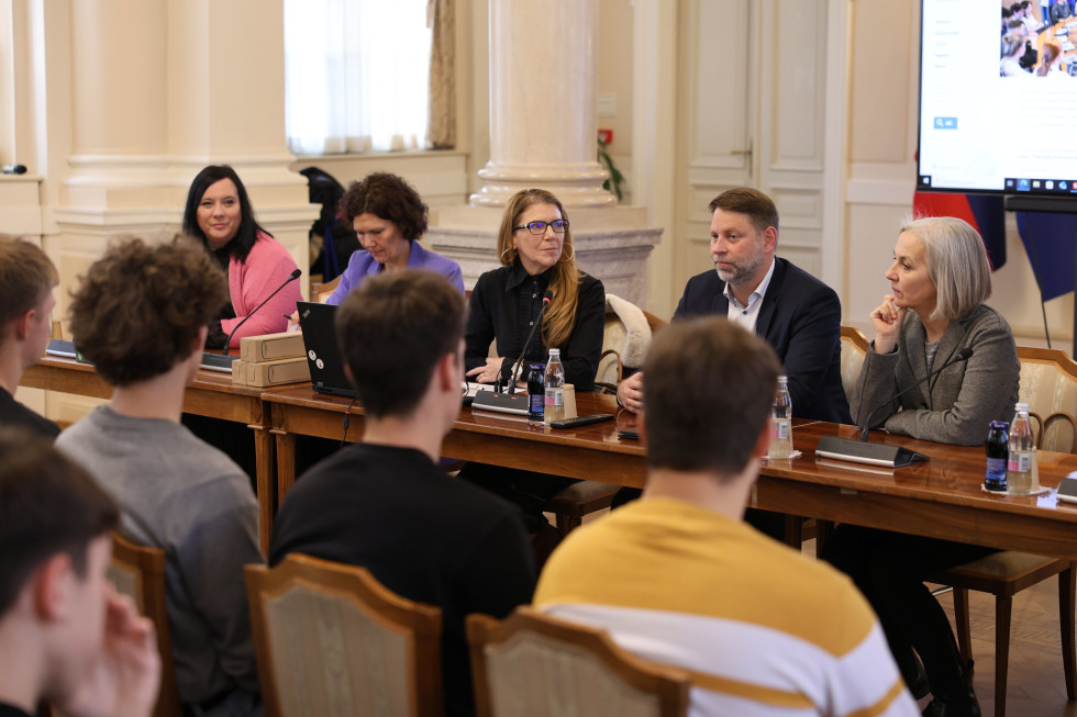 Štiri ženske in moški v formalnih oblačilih sedijo za mizo, pred njimi mikrofoni. 