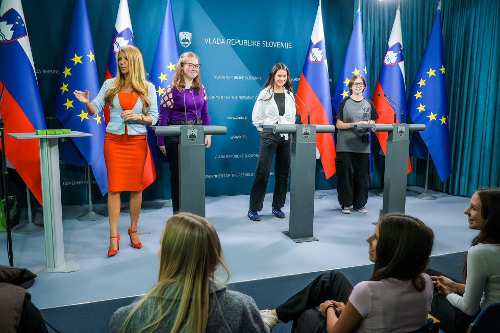 Tri mlajše ženske stojijo za govorniškim pultom v novinarskem središču, na levi strani ženska stoji in kaže z roko proti levi strani.