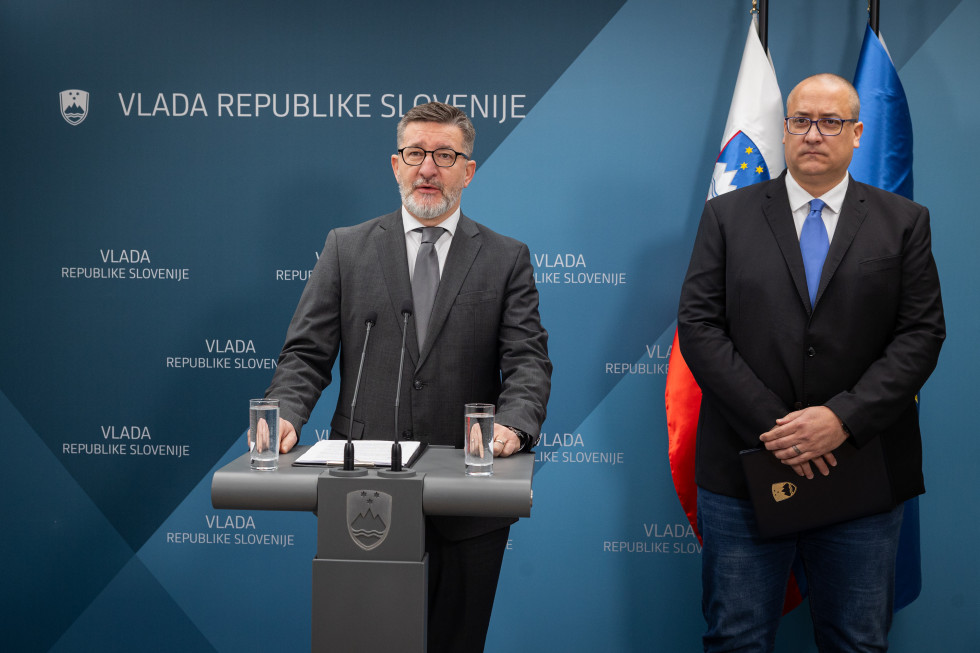 Branko Lobnikar stoji za govorniškim pultom, Jure Leben stoji na desni, za njim so zastave.