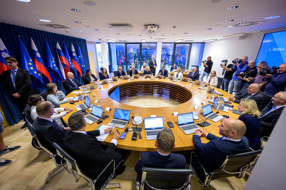 People seated around a round table with computers in front of them in a room with panoramic windows and Slovenian and EU flags.