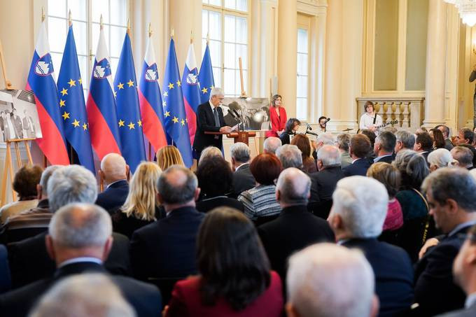 dr. Ciril Ribičič ob govorniškem pultu in sedeči gostje prireditve v veliki dvorani predsedniške palače