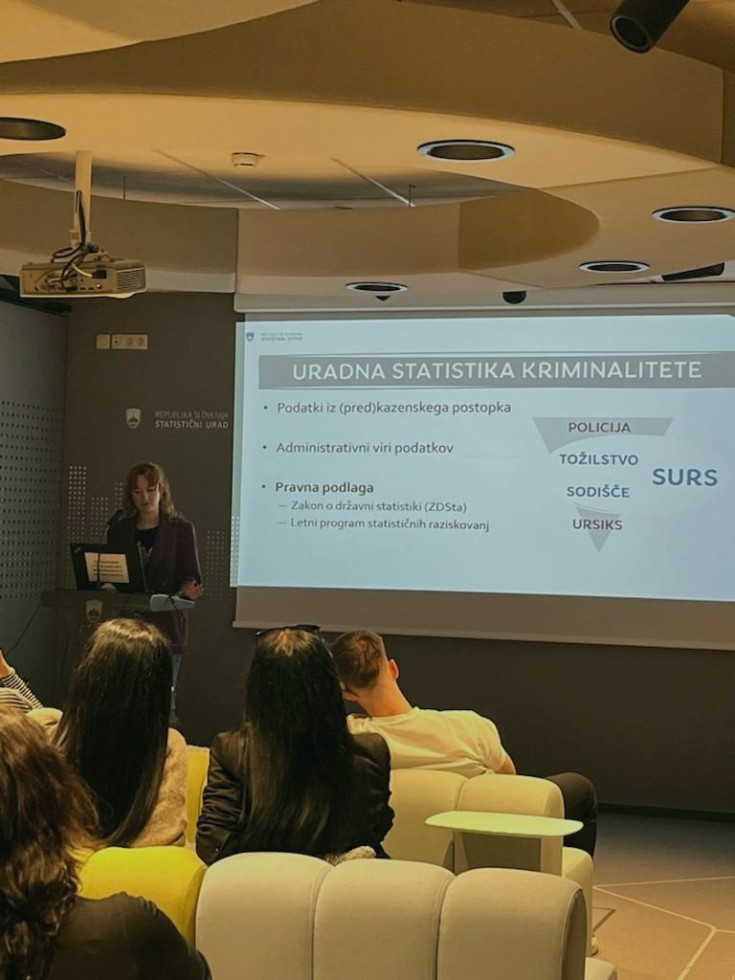 Strokovnjakinja za statistiko kriminalitete stoji za govorniškim pultom in predstavlja predstavitev na projekcijskem platnu v konferenčni sobi. Na platnu je naslov o uradni statistiki kriminalitete ter seznam virov podatkov in pravne podlage.