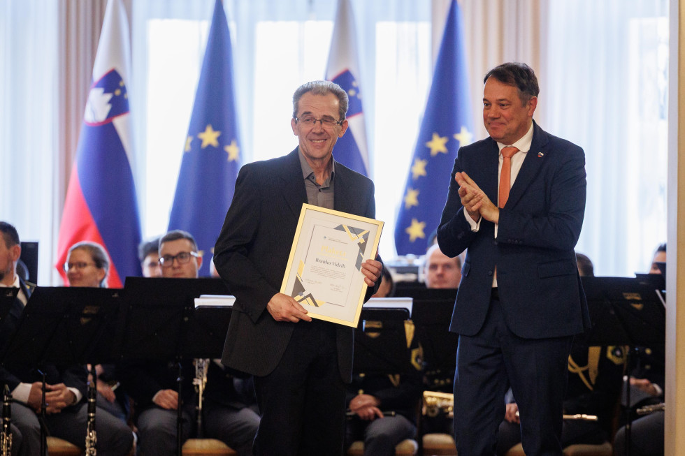 Moški s plaketo v roki. Drug moški mu ploska. Zadaj slovenske in evropske zastave.