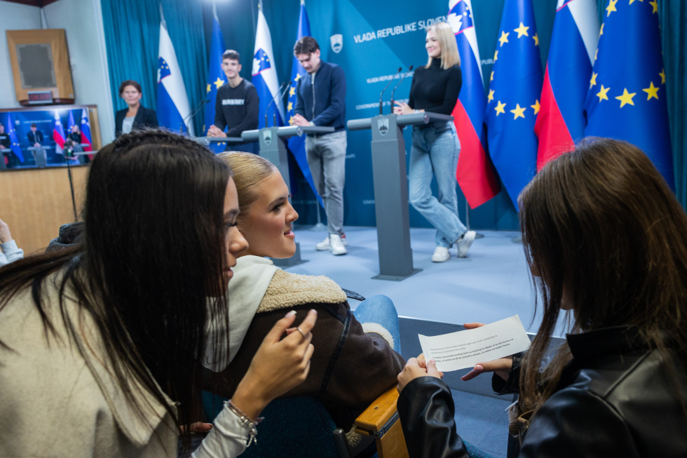 Tri mlada dekleta gledajo listek, v ozadju štirje ljudje na odru za govorniškimi pulti.