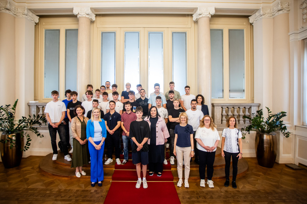 Večja skupina odraslih in otrok stoji v večji dvorani ter pozira za skupinsko fotografijo na stopnišču z rdečo preprogo.