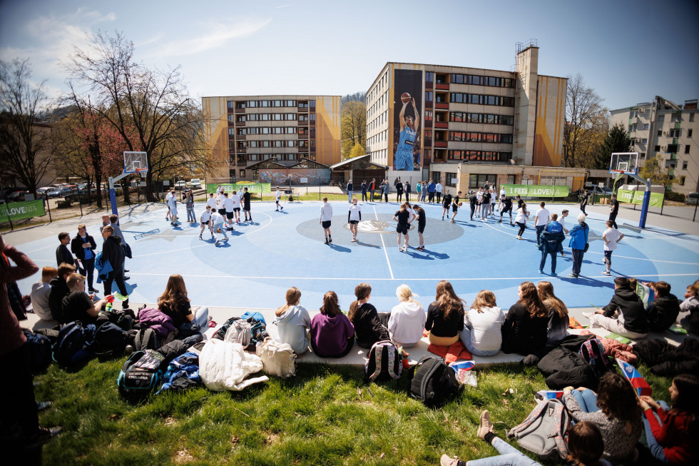 Otroci sedijo na travi in gledajo tekmo. Pogled na košarkarsko igrišče modre barve, poleg njega so dijaški domovi.