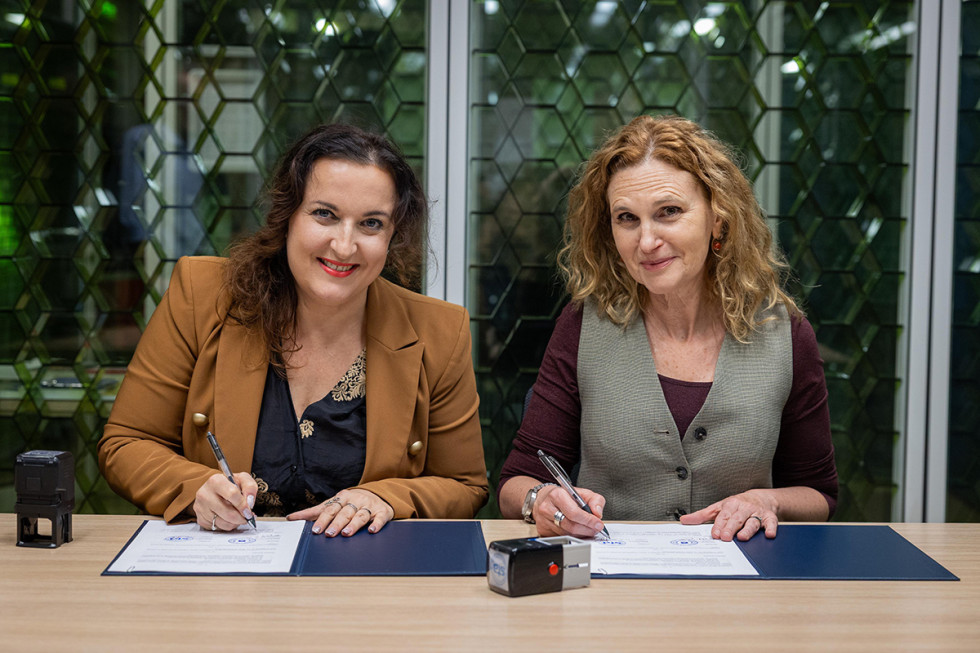 Two representatives of institutions are sitting at a table and signing documents in blue folders; a stamp is visible on the table, and in the background there is a glass wall.