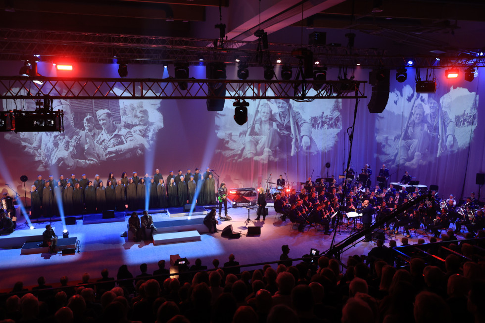 Igralci, pevski zbor in orkester na odru. V ozadju je projekcija starih fotografij iz 2. svetovne vojne. Občinstvo sedi v dvorani.