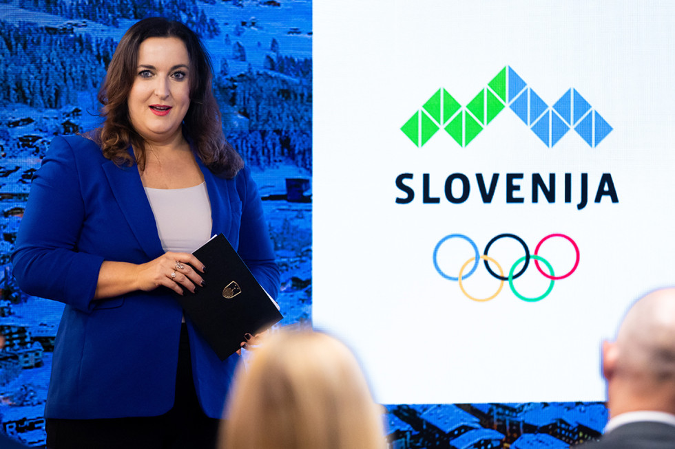 Oseba na odru pred občinstvom; v ozadju je prikazan logotip Olimpijskega komiteja Slovenije.