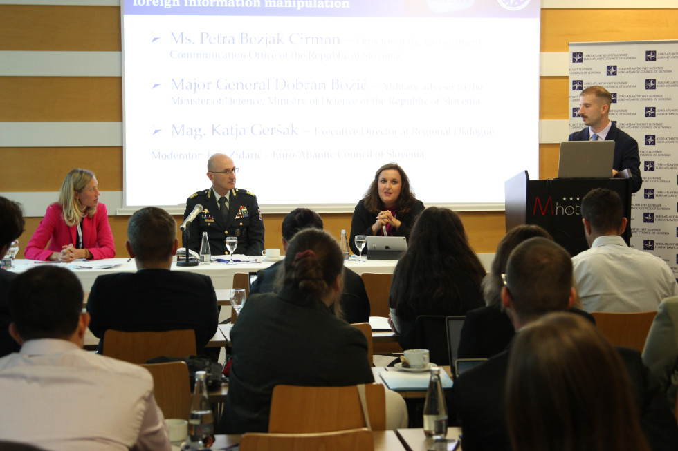 The Director of the Government Communication Office sits at a desk next to the other two speakers, speaking to students in the hall. Next to the table is a lectern with a moderator standing behind it.
