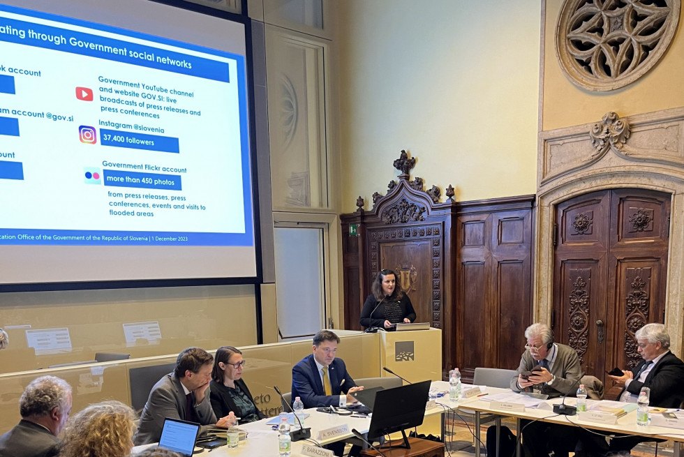 Petra Bezjak Cirman stands at the lectern. Participants are seated at a table. A presentation is displayed on the wall.
