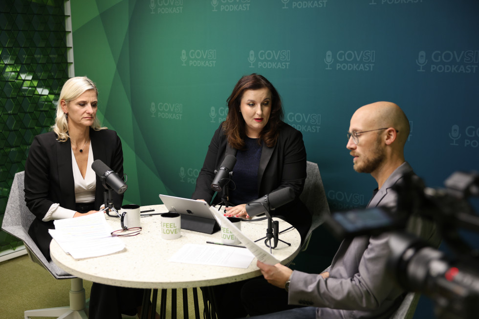Two women and a man sitting at a round table and talking in a green room.obi z zelenim ozadjem in se pogovarjajo.