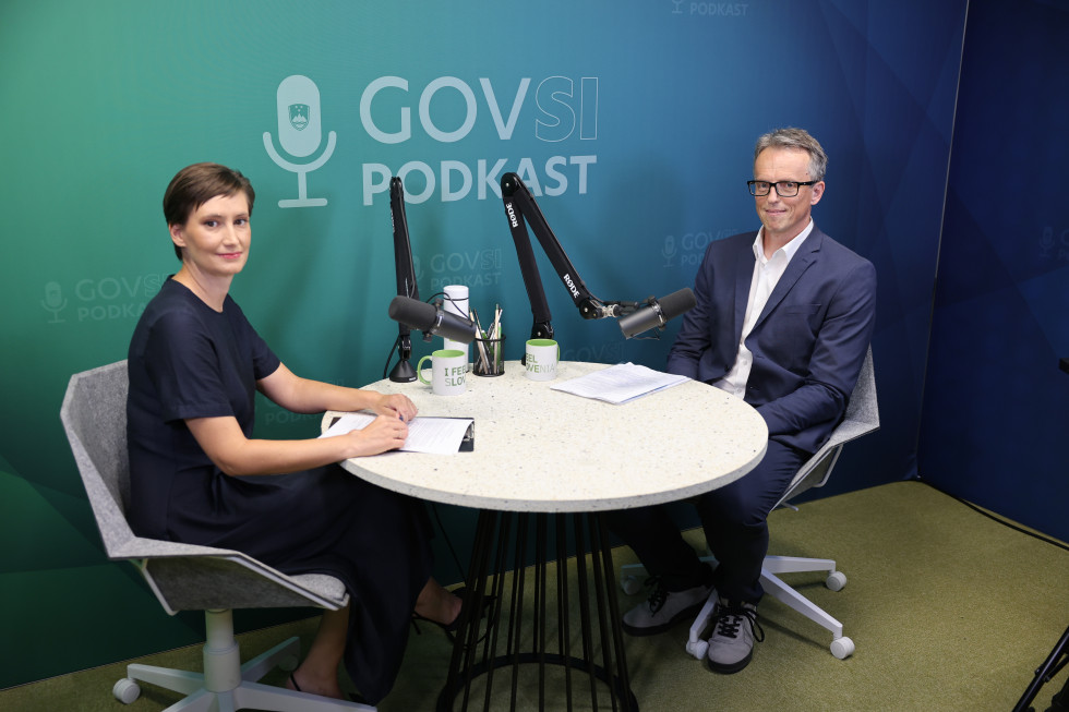 A woman and a man are seated at a round table with microphones, looking directly at the camera. Behind them, against a green background, is the text GOVSI Podcast.