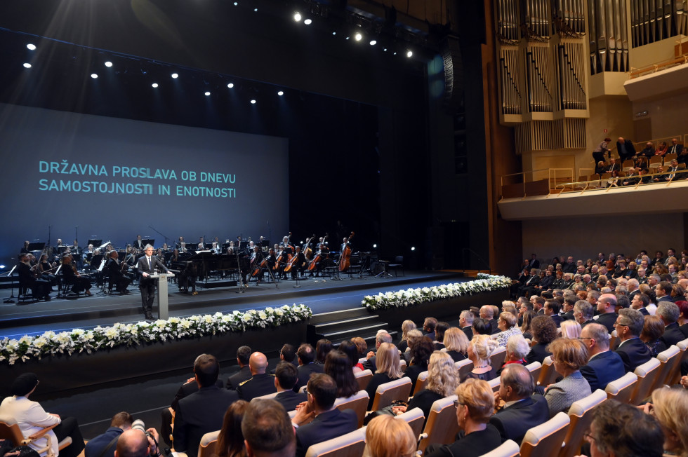 Orkester na odru, predsednik vlade Robert Golob stoji za govorniškim pultom, občinstvo sedi v dvorani. 