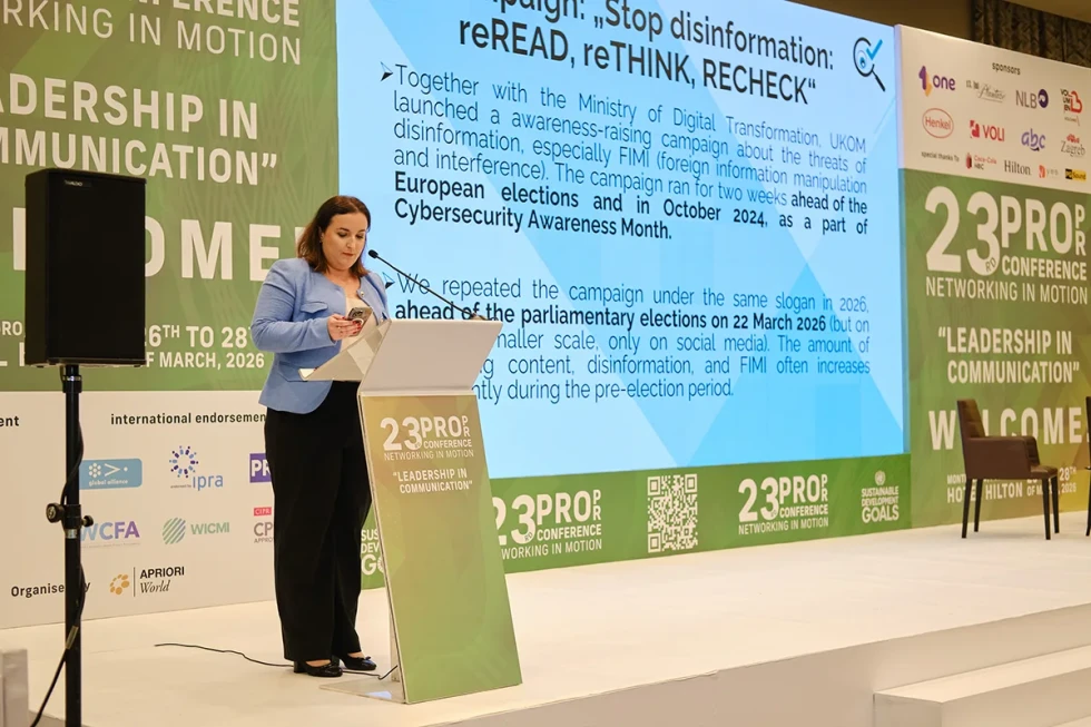 Govornica stoji za govorniškim pultom, v ozadju je projekcija predstavitve o kampanji proti dezinformacijam.