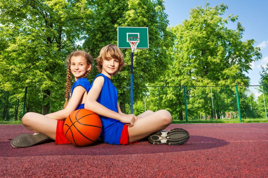 Deklica in deček sedita na košarkarskem igrišču. Poleg imata košarkarsko žogo.