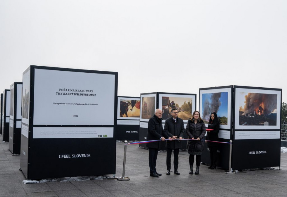 Janko Boštjančič, Marjan Šarec in Petra Bezjak Cirman režejo trak ob otvoritvi razstave. V ozadju kubusi s fotografijami razstave.