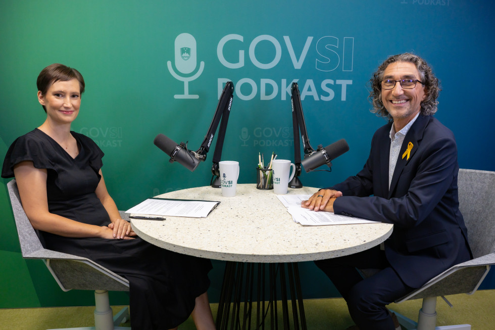 A woman and a man sit at a round table with microphones in a GOVSI PODKAST studio, smiling; papers and mugs on the table.
