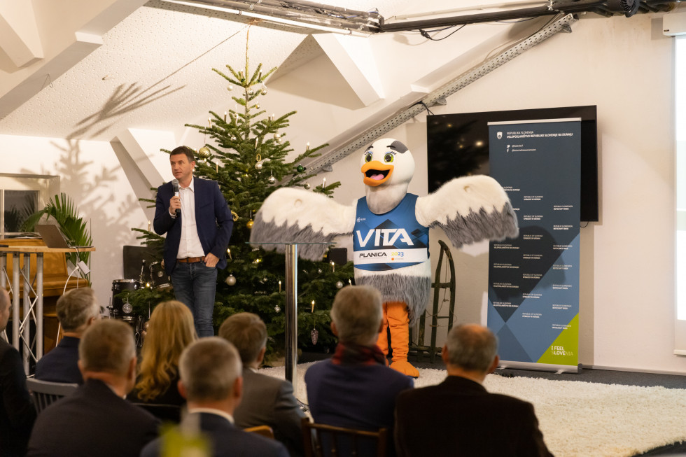 A hall with an audience, a speaker and the mascot Vita stand on the stage.