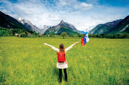 Dekle, obrnjena s hrbtom, s stegnjenimi rokami, drži slovensko zastavo. Pogled na travnik in gore.