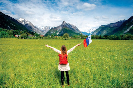 A girl, with her back turned and her arms outstretched, holds the Slovenian flag. View of the meadow and mountains.