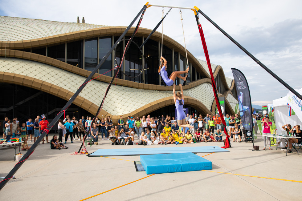 People are sitting in front of the Arena Stožice, two acrobats are performing a circus performance.