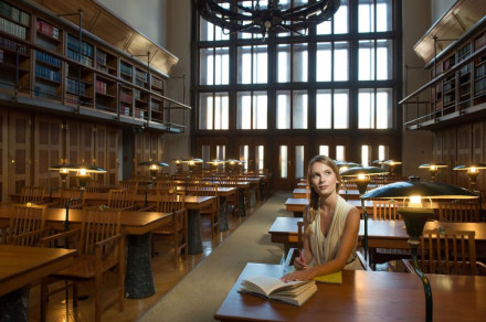 Dekle sedi za mizo v Narodni univerzitetni knjižnici. Na mizi ima knjigo in zvezek za zapiske.