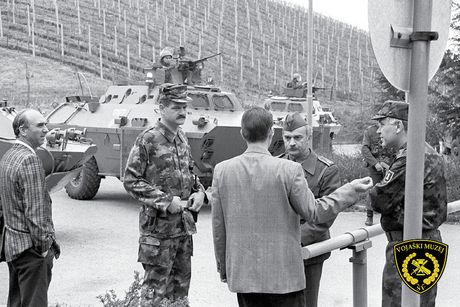 Ljudje, med njimi vojak, se pogovarjajo na cesti. V ozadju tank.