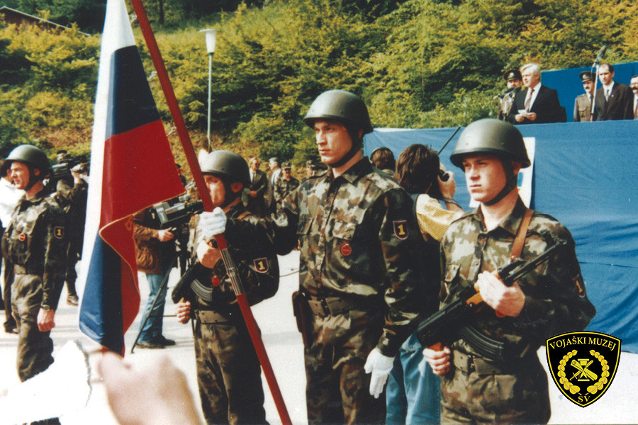 V ospredju stojijo zastavonoše s slovensko zastavo. V ozadju govorniški oder. Milan Kučan govori, poleg njega stoji Janez Janša.