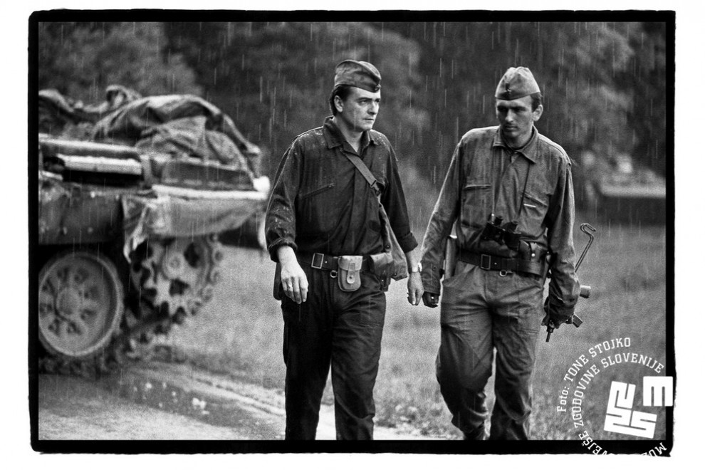 Vojaka JLA hodita v dežju, v ozadju tank. 