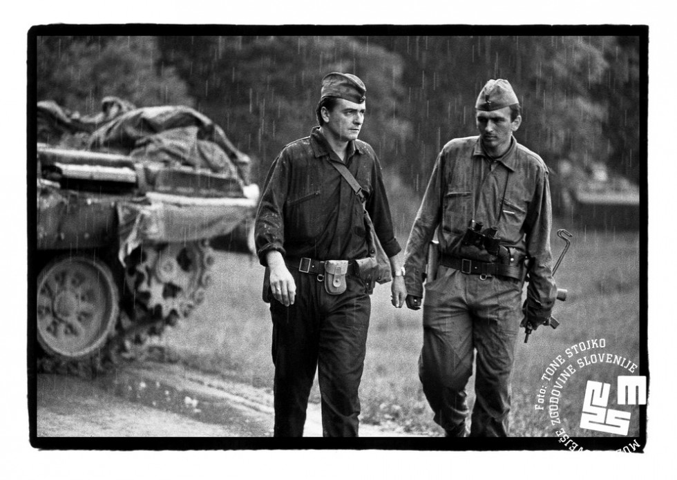 Vojaka JLA hodita v dežju, v ozadju tank. 