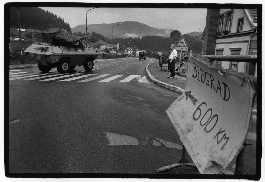 Tank na cesti, moški stoji na pločniku. Ob cesti je nekdo postavil tablo, na kateri piše Beograd 600 km.