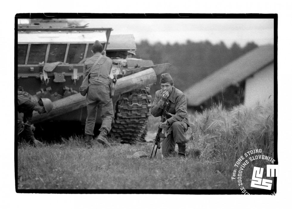 Trije vojaki jugoslovanske armade ob tanku. Eden čepi s puško v roki in kadi cigareto, druga dva sta pri tanku.