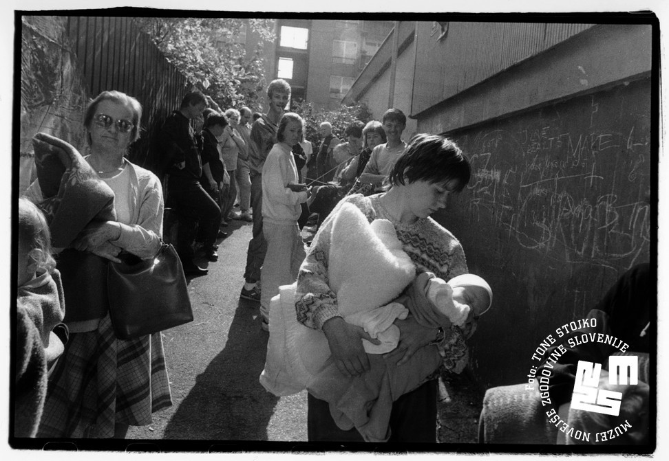 Blokovsko naselje, ljudje stojijo v vrsti pred zakloniščem, v ospredju mati z dojenčkom in starejša gospa, v ozadju najstniki.