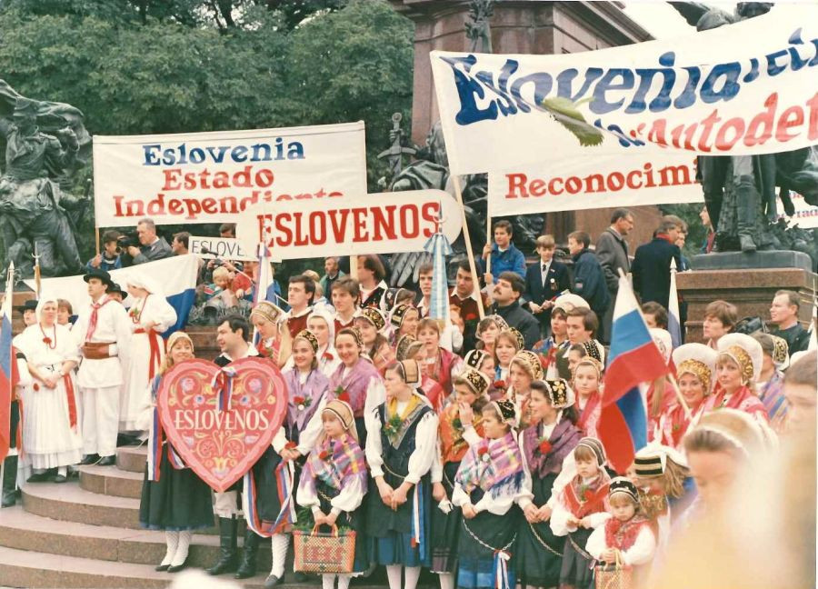 Ljudje v slovenskih narodnih nošah stojijo s plakati in panoji z napisi v španščini za neodvisno in samostojno Slovenijo.